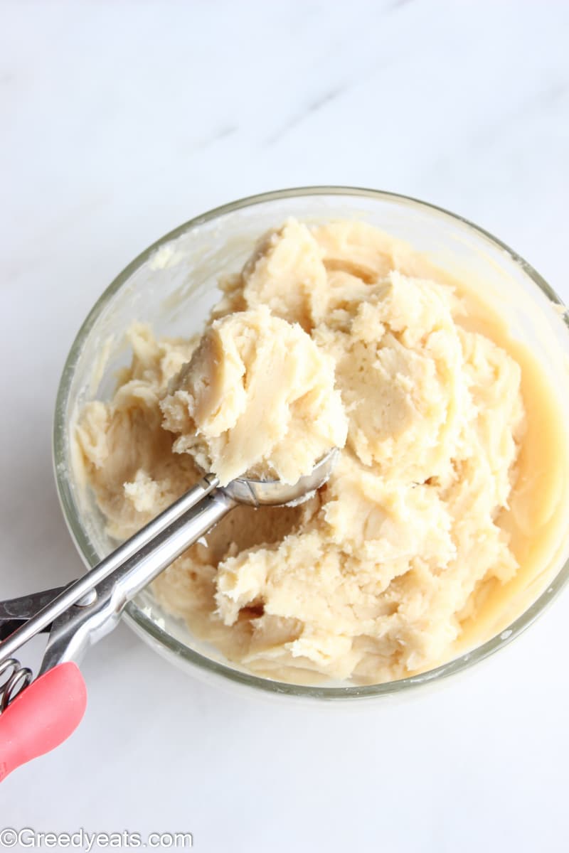 Chilled cream cheese cookie dough in a glass bowl with a cookie scoop in it.