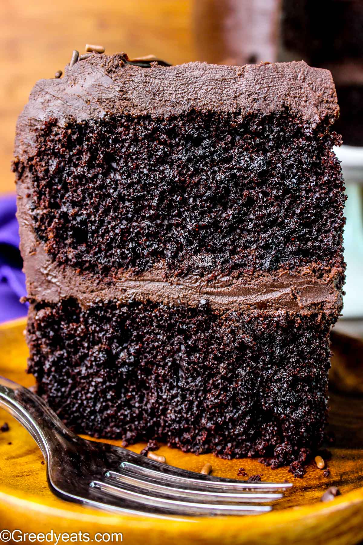 Moist and soft Chocolate Cake slice covered in fudge frosting on a wooden plate.