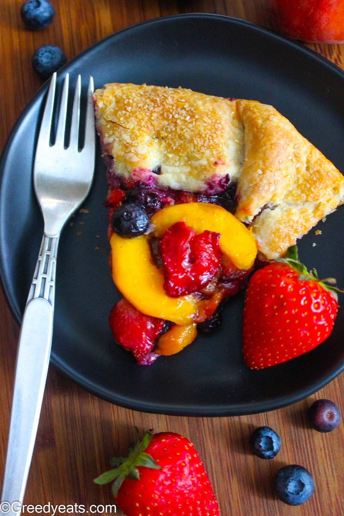 A slice of flaky Galette on black plate with peach and berries filling.