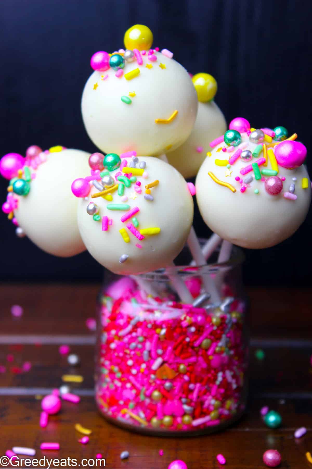 Homemade Cape Pops topped with sprinkles, held on lollipop sticks kept in a jar of sprinkles.