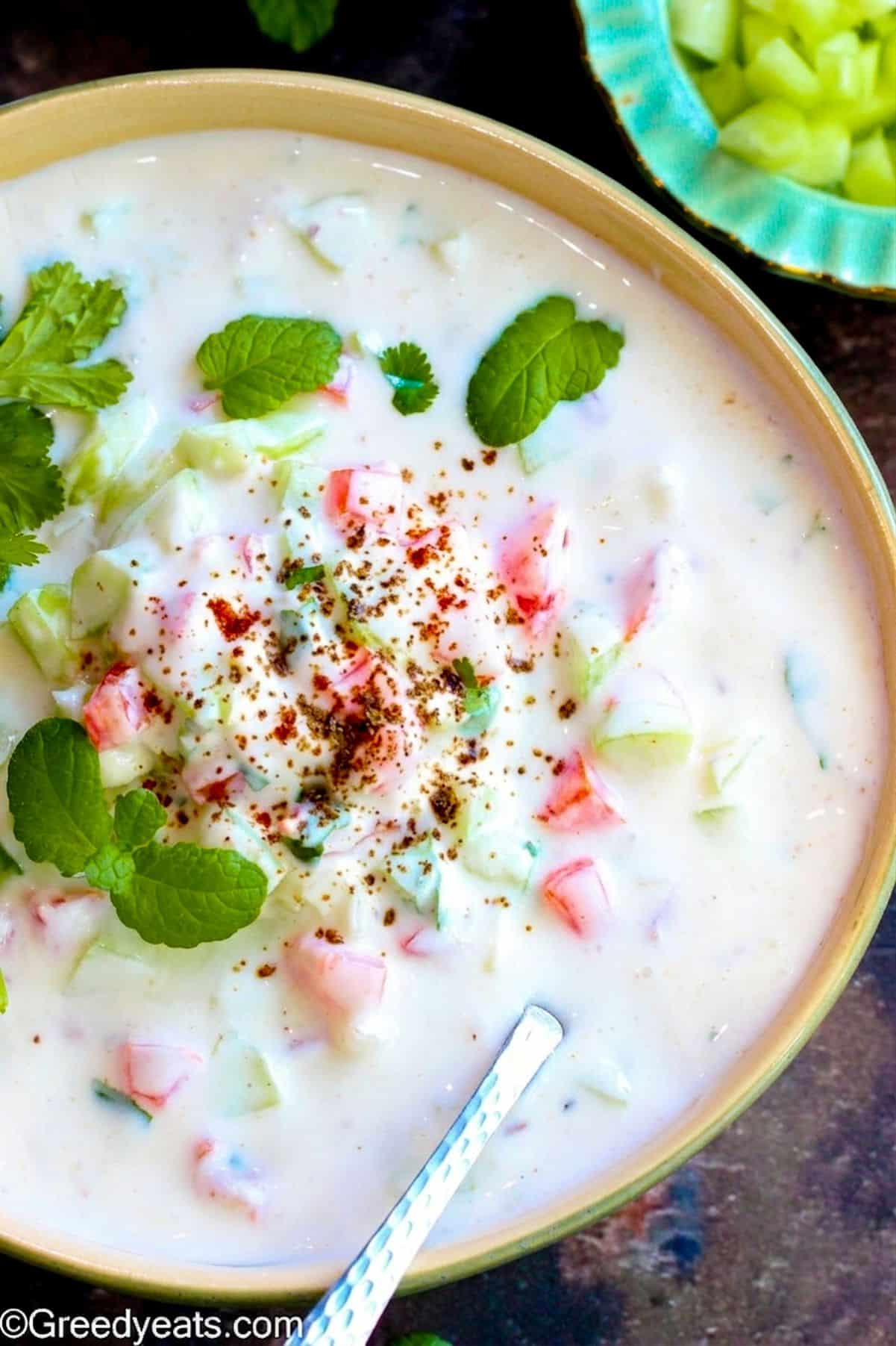 Raita recipe made with yogurt, herbs, spices and veggies served in a bowl.