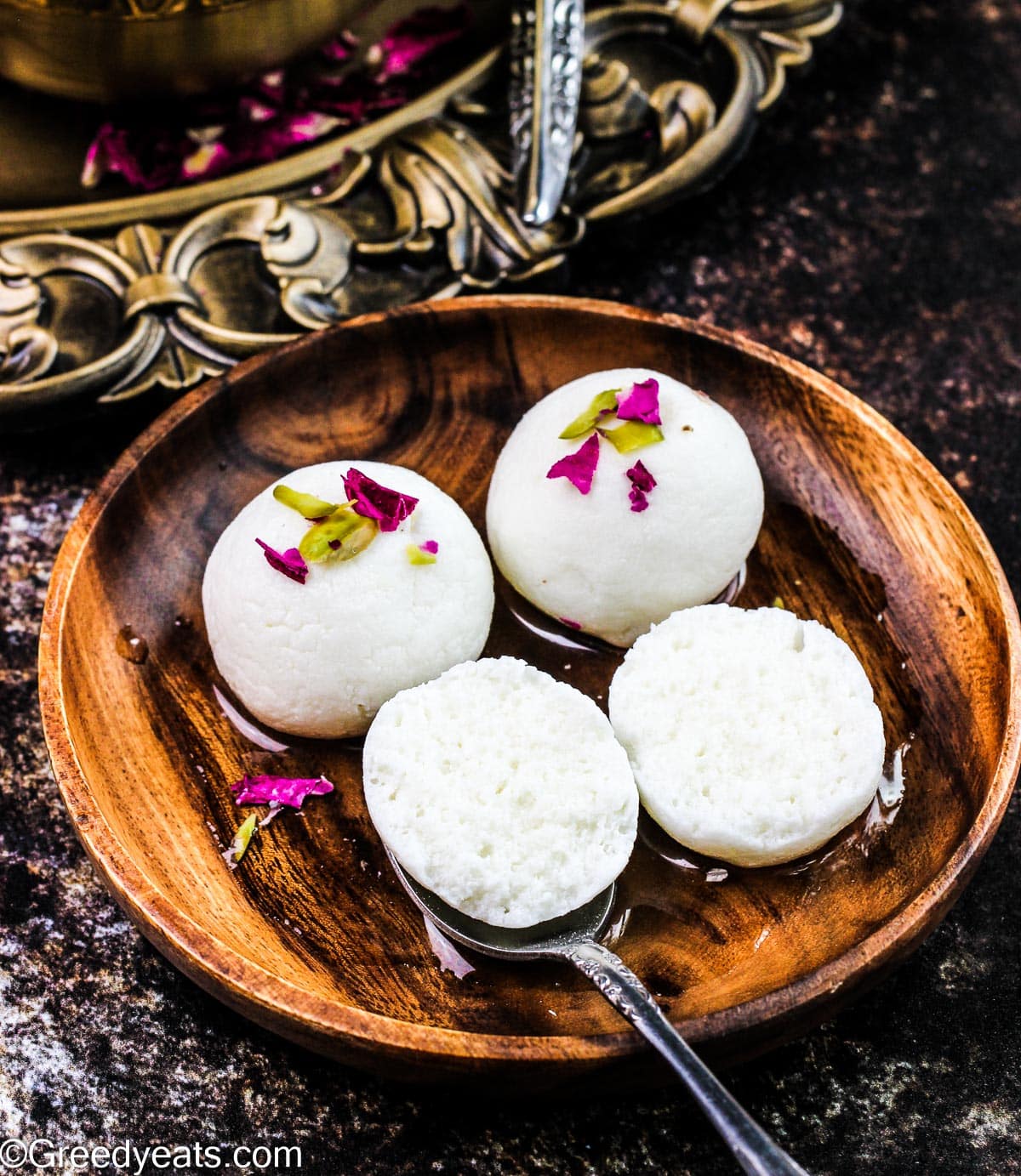 Soft and spongy texture of rasgulla is shown in the image served with rose petals and pistachios.