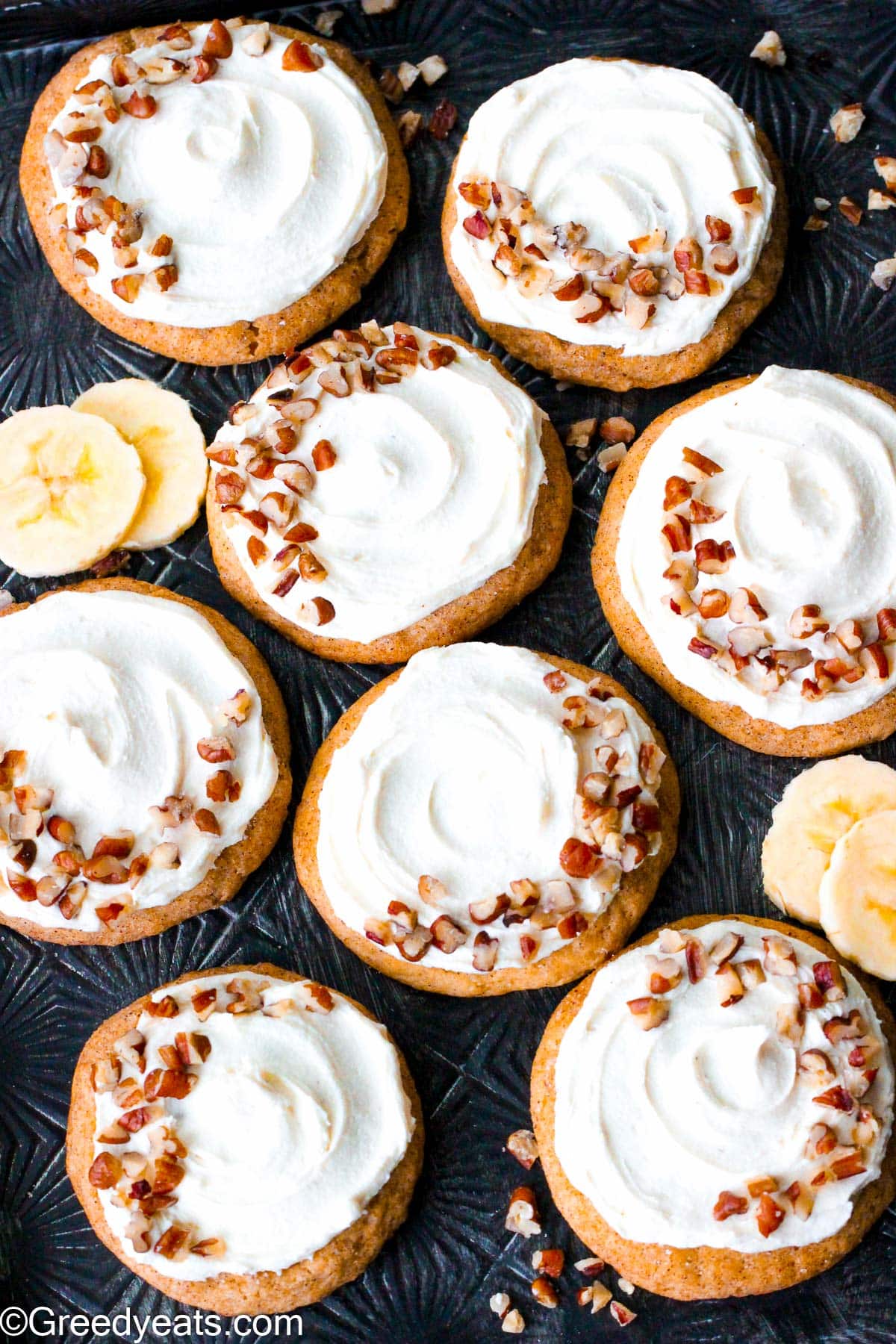 Banana cookies served on a baking tray topped with cream cheese frosting and chopped pecans.